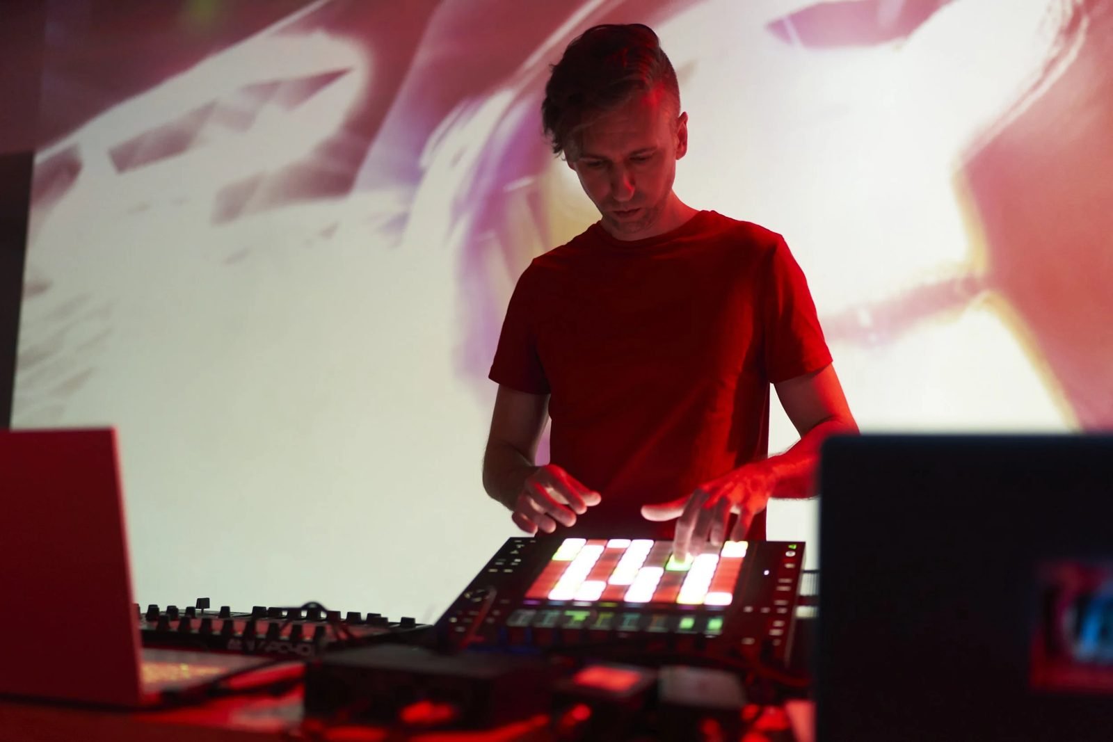 A performer at a laptop lit red, projected imagery wrapping the wall behind