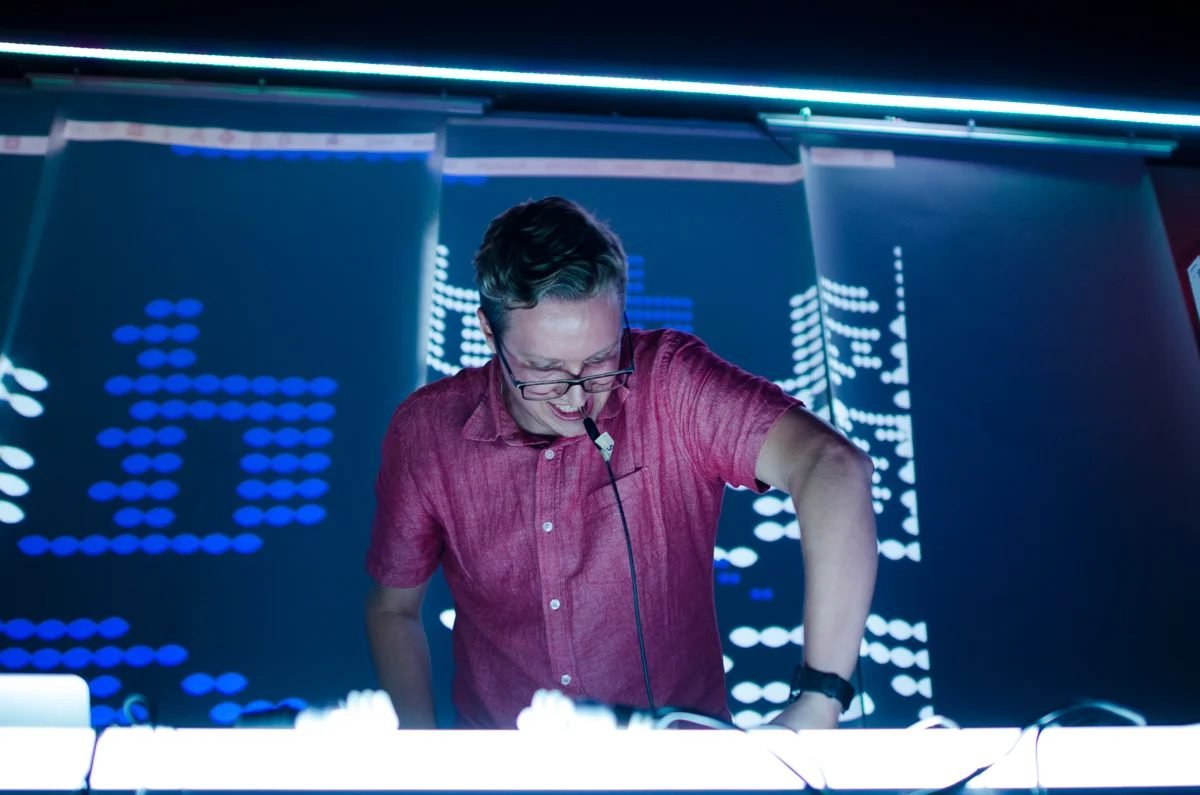 A performer in a red shirt behind a console, blue pixel-block projections behind