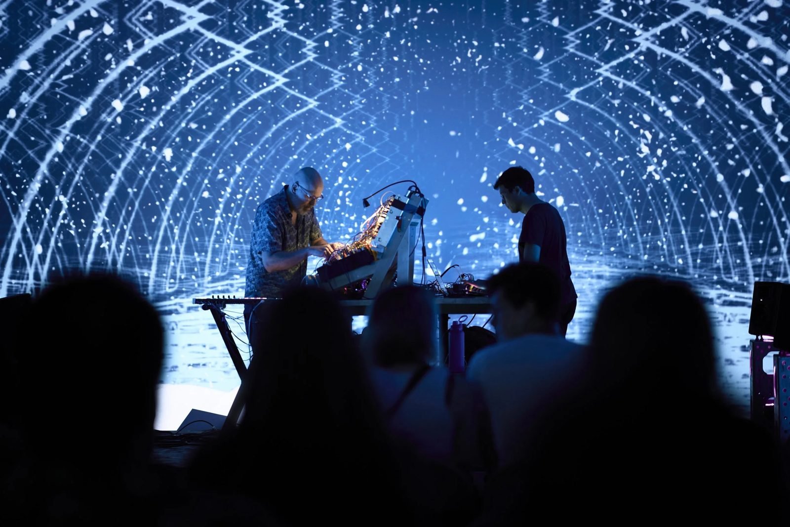 A crowd in silhouette facing two performers under a dome of projected white light grids at dadaLab