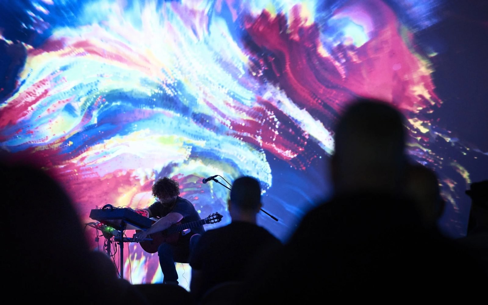 A seated guitarist performs in front of a wall of swirling pink, blue and yellow projected light