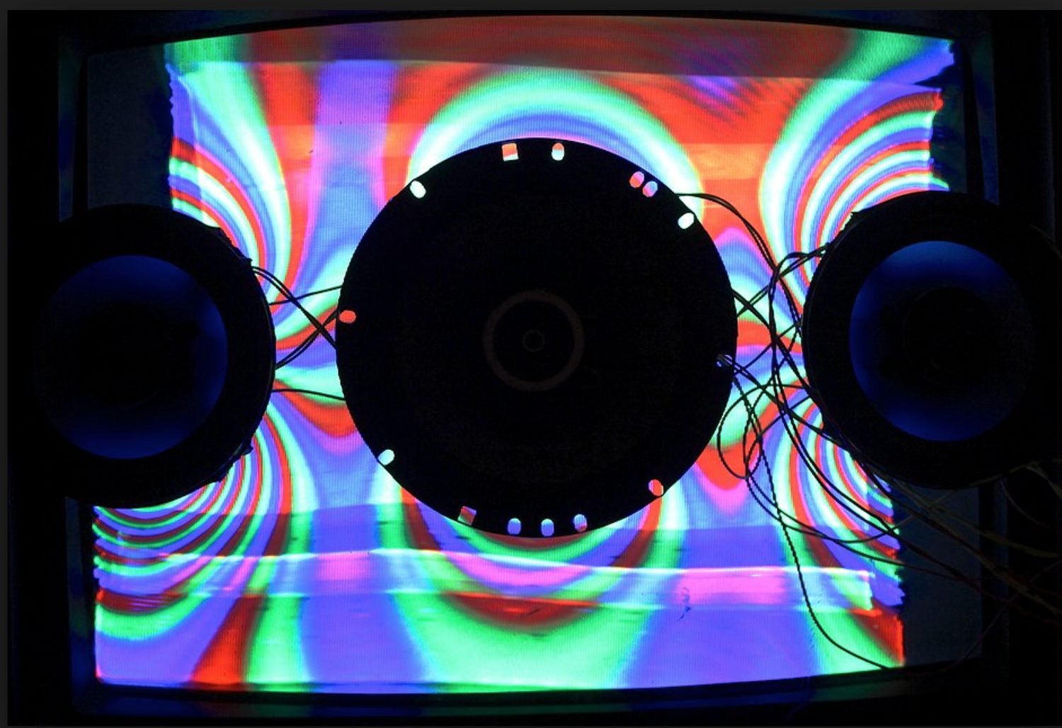 Two speakers flanking a round subwoofer in front of a wall of rainbow concentric projection